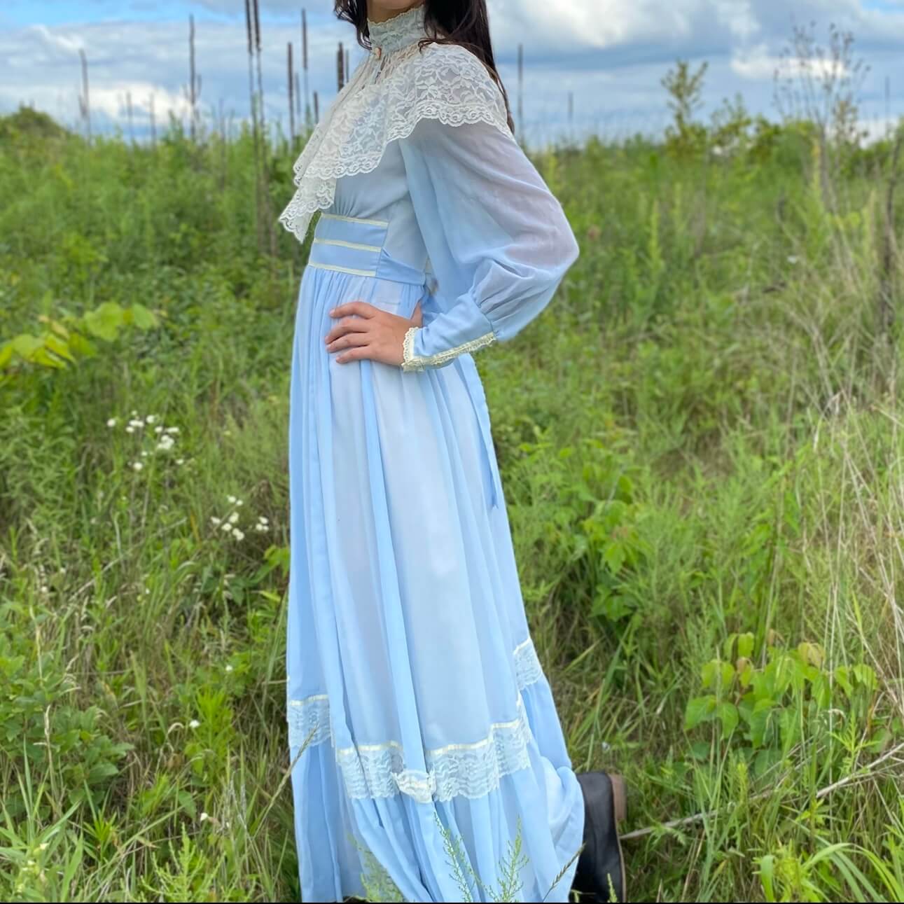 model wearing a blue cotton long dress from the 70s with a white lace high neck collar in front of green grass