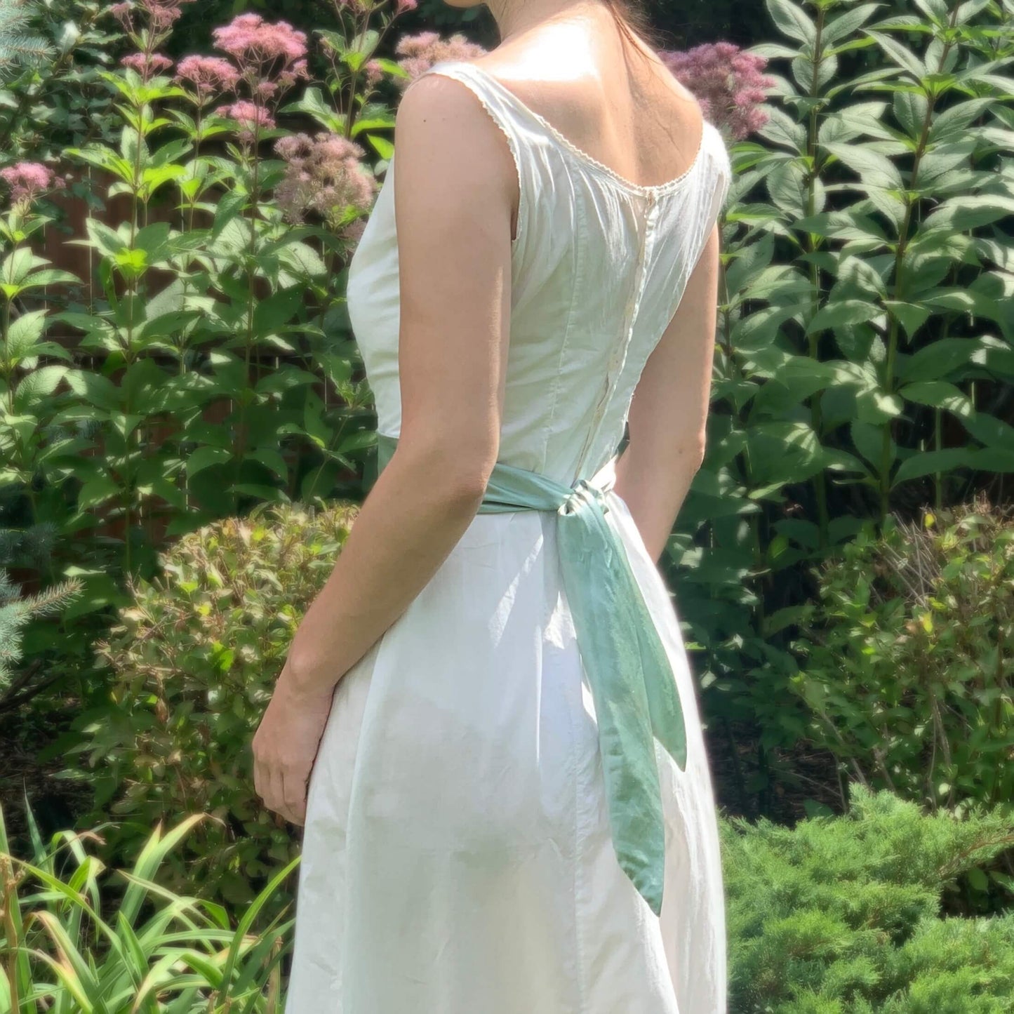 back view of an edwardian chemise dress with a blue waist sash worn on a girl in front of flowers