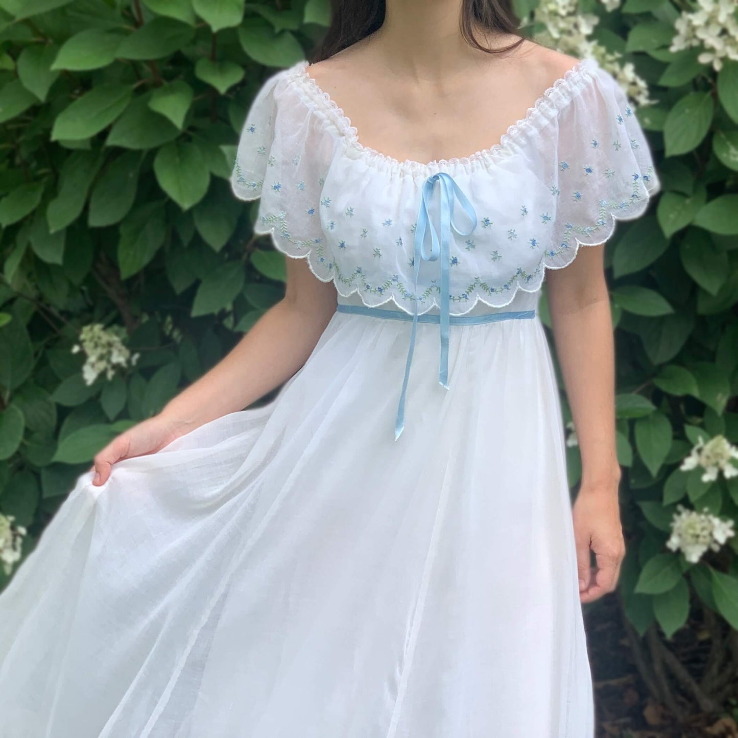 vintage gunne sax wedding dress with blue embroidery and a bow on a model in front of a green bush
