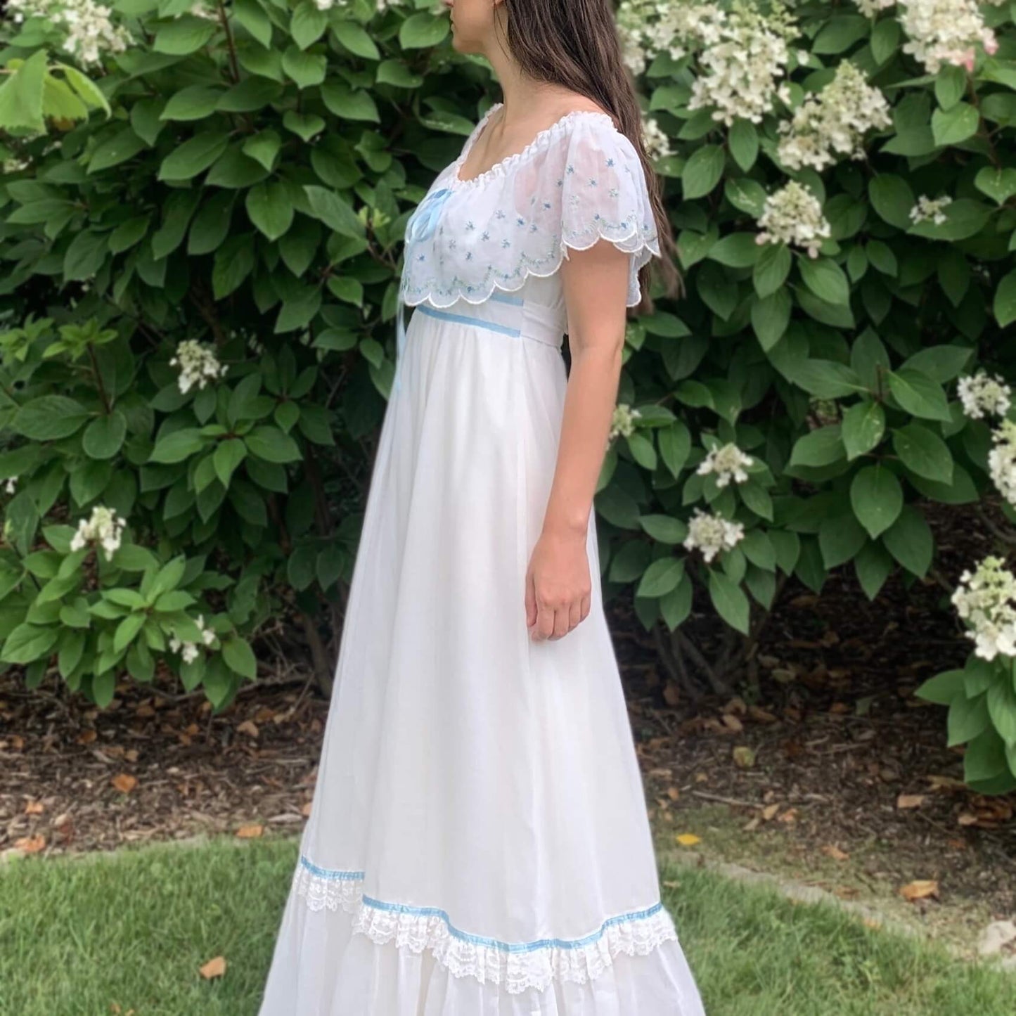 side view of a girl wearing a maxi dress standing on grass in front of a flower bush